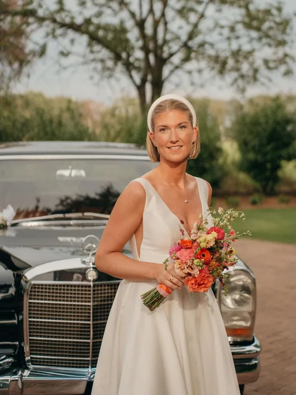 Braut mit Sleek-Bob, Haarreif und buntem Brautstrauß vor einem Mercedes-Oldtimer
